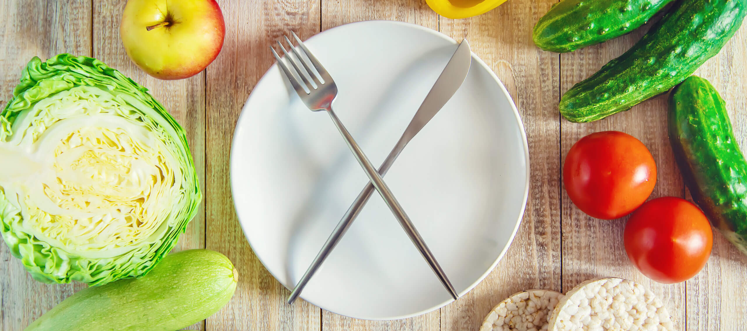An empty plate with utensils forming an “X” at the center, surrounded by a scattered arrangement of fruits and vegetables in the background.